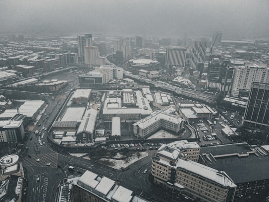 An aerial view of a cityscape in winter, with buildings, roads, and parking lots covered in a light layer of snow. The atmosphere is gray and overcast, suggesting cold weather. High-rise buildings are visible in the background, with several main roads intersecting through the area.