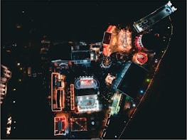 An aerial view of a brightly lit amusement park at night, showcasing various attractions including a ferris wheel, carnival games, and pathways outlined with festive lights. The park is bustling with activity, with colorful tents and structures creating a vibrant contrast against the darkness.