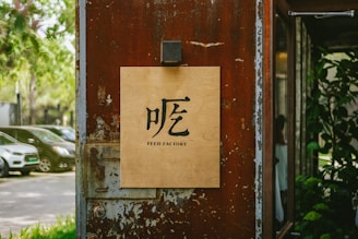 A wooden sign with the text 'FEED FACTORY' and a Chinese character is mounted on a weathered, rust-colored metal wall. The background features parked cars and green trees, creating a blend of urban and natural elements.