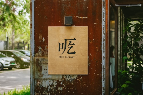 A wooden sign with the text 'FEED FACTORY' and a Chinese character is mounted on a weathered, rust-colored metal wall. The background features parked cars and green trees, creating a blend of urban and natural elements.