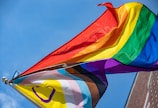 Two flags with vibrant rainbow colors are waving against a clear blue sky. The flags include symbols and additional colors representing diversity and inclusion.