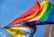 Two flags with vibrant rainbow colors are waving against a clear blue sky. The flags include symbols and additional colors representing diversity and inclusion.