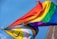 Two flags with vibrant rainbow colors are waving against a clear blue sky. The flags include symbols and additional colors representing diversity and inclusion.