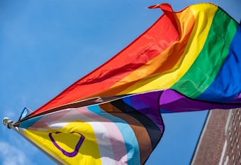Two flags with vibrant rainbow colors are waving against a clear blue sky. The flags include symbols and additional colors representing diversity and inclusion.