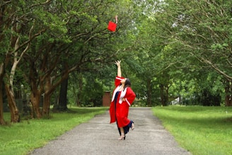 A person wearing a red graduation gown and cap is standing on a tree-lined pathway, joyfully throwing the cap into the air. The surroundings are lush with greenery, and the pathway leads to a more tree-dense area in the background.