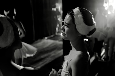 Ballet dancers are preparing backstage, with one prominently featured woman in a stage costume and headpiece. Soft lighting highlights their expressions of calm concentration.