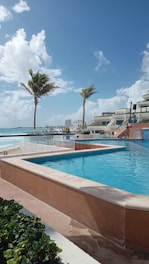 A luxurious outdoor pool area overlooking a beach and ocean, with palm trees swaying gently in the breeze. A few buildings are visible in the background beneath a clear blue sky spotted with fluffy white clouds. The area is tranquil and inviting.
