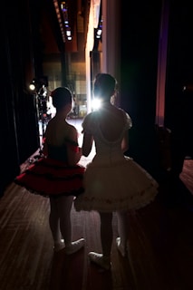 Two young ballerinas stand backstage, dressed in elaborate tutus and ballet shoes, facing a brightly lit stage. The setting conveys anticipation as they wait to perform. The lighting creates a dramatic silhouette effect, emphasizing their poised posture and elegant costumes.