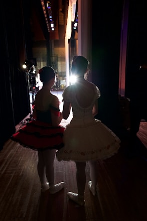 Two young ballerinas stand backstage, dressed in elaborate tutus and ballet shoes, facing a brightly lit stage. The setting conveys anticipation as they wait to perform. The lighting creates a dramatic silhouette effect, emphasizing their poised posture and elegant costumes.