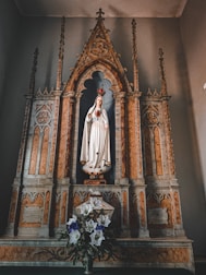 A detailed altar with ornate decorations, featuring a statue of a woman in a white robe, adorned with a crown and holding a heart symbol. The altar is designed with intricate carvings and marble-like features. In front of the statue, there is a bouquet of white and purple flowers.