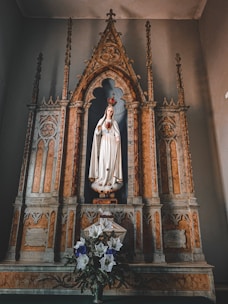 A detailed altar with ornate decorations, featuring a statue of a woman in a white robe, adorned with a crown and holding a heart symbol. The altar is designed with intricate carvings and marble-like features. In front of the statue, there is a bouquet of white and purple flowers.