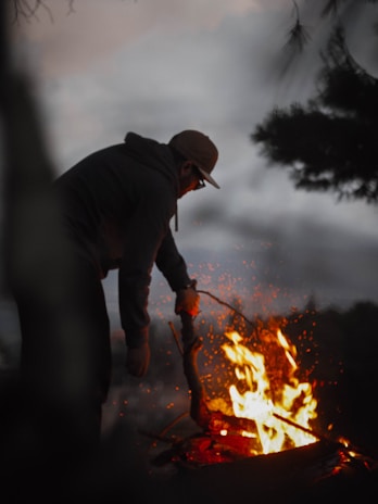 A person in a hoodie and cap tends to a campfire with a stick, surrounded by a dark, forested environment. The flames are vivid and illuminate the scene with glowing sparks, contrasting against the dim background.