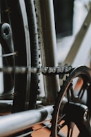 Close-up view of a bicycle's drivetrain, featuring the chain, gear, and sections of the wheel. The metal components show signs of wear and use, with the chain fitted securely on the gear.