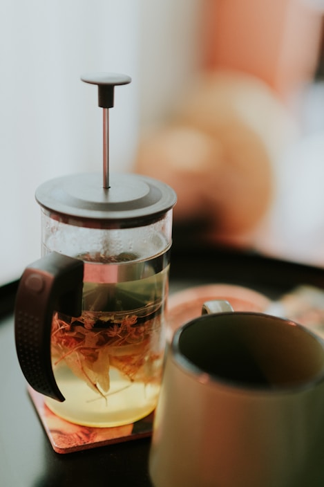 A French press filled with herbal tea sits on a coaster next to an empty ceramic mug. The background is softly blurred, suggesting a cozy indoor setting.