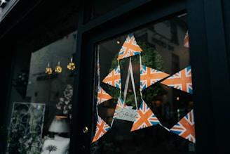 A window display features a decorative arrangement with triangular Union Jack banners forming a star pattern, framing a green wreath. Below the wreath, a small sign hangs with the word 'open' written on it. Inside the shop, various decor items and plants are visible.