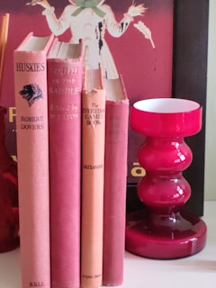 Several vintage books with red and pink covers are neatly arranged vertically on a white surface. Next to the books is a glossy red candle holder or decorative vase with a modern design. In the background, there is part of a poster featuring an individual holding what appears to be stalks of wheat.