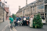 A group of people dressed in festive, colorful attire, some holding musical instruments like drums and tambourines, standing on a street in front of a historic stone building. Some are adorned with flowers and greenery, suggesting a celebration or festival. The scene includes a diverse mix of individuals, including one person with a walker decorated with plants.