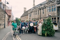 A group of people dressed in festive, colorful attire, some holding musical instruments like drums and tambourines, standing on a street in front of a historic stone building. Some are adorned with flowers and greenery, suggesting a celebration or festival. The scene includes a diverse mix of individuals, including one person with a walker decorated with plants.