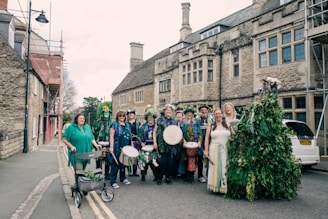 A group of people dressed in festive, colorful attire, some holding musical instruments like drums and tambourines, standing on a street in front of a historic stone building. Some are adorned with flowers and greenery, suggesting a celebration or festival. The scene includes a diverse mix of individuals, including one person with a walker decorated with plants.