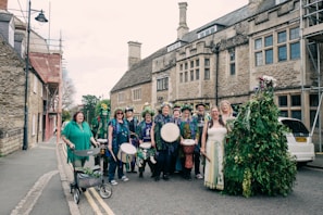 A group of people dressed in festive, colorful attire, some holding musical instruments like drums and tambourines, standing on a street in front of a historic stone building. Some are adorned with flowers and greenery, suggesting a celebration or festival. The scene includes a diverse mix of individuals, including one person with a walker decorated with plants.