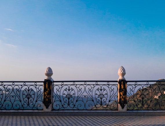 An ornate metal railing with intricate patterns stands on a tiled floor. The tall, decorative pillars of the railing frame a scenic view of distant mountains and a clear blue sky with subtle clouds.