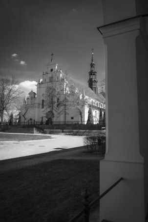 A large, ornate church with a tall, intricate spire stands prominently under a partly cloudy sky. The foreground includes a paved pathway and manicured gardens with trees shedding leaves, conveying a serene atmosphere. The architecture features classical design elements, with arched windows and decorative moldings.