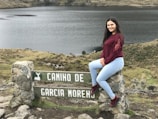 A young woman with long dark hair, wearing a maroon top and light blue jeans, sits on a stone sign with the text 'CAMINO DE GARCIA MORENO'. The landscape features a serene lake and grassy hillsides, creating a peaceful natural setting.