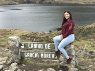 A young woman with long dark hair, wearing a maroon top and light blue jeans, sits on a stone sign with the text 'CAMINO DE GARCIA MORENO'. The landscape features a serene lake and grassy hillsides, creating a peaceful natural setting.