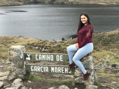 A young woman with long dark hair, wearing a maroon top and light blue jeans, sits on a stone sign with the text 'CAMINO DE GARCIA MORENO'. The landscape features a serene lake and grassy hillsides, creating a peaceful natural setting.