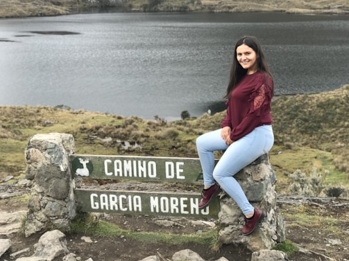 A young woman with long dark hair, wearing a maroon top and light blue jeans, sits on a stone sign with the text 'CAMINO DE GARCIA MORENO'. The landscape features a serene lake and grassy hillsides, creating a peaceful natural setting.