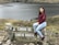 A young woman with long dark hair, wearing a maroon top and light blue jeans, sits on a stone sign with the text 'CAMINO DE GARCIA MORENO'. The landscape features a serene lake and grassy hillsides, creating a peaceful natural setting.