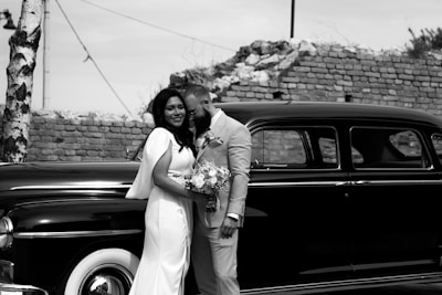 The bride and groom standing regally beside a vintage wedding car decorated with vibrant red flowers under soft, romantic lighting.
