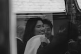 A black and white photograph captures a joyful moment inside a vehicle. The focus is on a smiling person whose expression conveys happiness and warmth. Additional figures are faintly visible in the background, adding depth to the scene. The overall mood is intimate and candid.