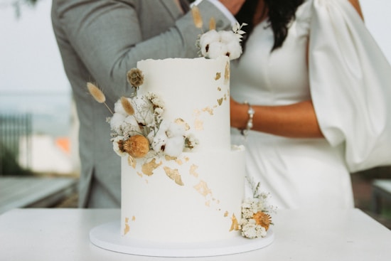 A two-tiered white cake adorned with gold leaf accents and decorated with dried flowers and cotton. In the background, a couple is present, partially visible, with the woman holding a pearl bracelet.
