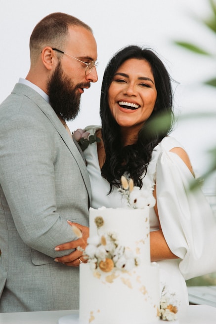 A couple stands close together, looking happy and celebratory. The woman is laughing joyfully and wears a white dress with floral highlights, while the man, dressed in a gray suit, holds her affectionately. In front of them is a white-tiered wedding cake decorated with flowers. The setting appears to be bright and possibly outdoors.