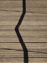 A wooden deck surface with a prominent zigzag shadow cast across it. Long, straight planks create a lined texture, and some small twigs or debris are scattered randomly.