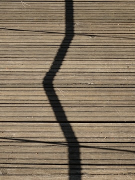 A wooden deck surface with a prominent zigzag shadow cast across it. Long, straight planks create a lined texture, and some small twigs or debris are scattered randomly.