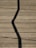 A wooden deck surface with a prominent zigzag shadow cast across it. Long, straight planks create a lined texture, and some small twigs or debris are scattered randomly.