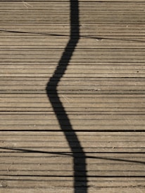 A wooden deck surface with a prominent zigzag shadow cast across it. Long, straight planks create a lined texture, and some small twigs or debris are scattered randomly.