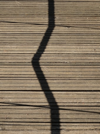 A wooden deck surface with a prominent zigzag shadow cast across it. Long, straight planks create a lined texture, and some small twigs or debris are scattered randomly.
