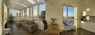A modern, well-lit living space with large windows offering a panoramic city view. The room features a gray sofa, a decorated Christmas tree, a wooden dining table with flowers and fruit, and a kitchenette in the corner. The floor is wooden, and the space appears open and airy.