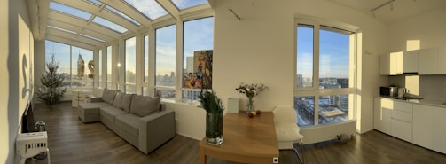 A modern, well-lit living space with large windows offering a panoramic city view. The room features a gray sofa, a decorated Christmas tree, a wooden dining table with flowers and fruit, and a kitchenette in the corner. The floor is wooden, and the space appears open and airy.