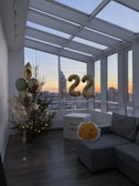 A modern indoor space with large windows offering a cityscape view during sunset. Decorated for a celebration, with a Christmas tree adorned with lights, a bouquet of yellow flowers, gold balloons, and a box labeled 'happy birthday.' Metallic number balloons displaying '22' are visible.