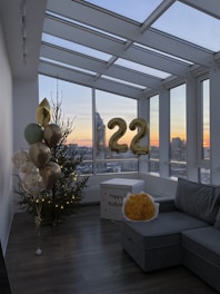 A modern indoor space with large windows offering a cityscape view during sunset. Decorated for a celebration, with a Christmas tree adorned with lights, a bouquet of yellow flowers, gold balloons, and a box labeled 'happy birthday.' Metallic number balloons displaying '22' are visible.