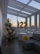 A modern indoor space with large windows offering a cityscape view during sunset. Decorated for a celebration, with a Christmas tree adorned with lights, a bouquet of yellow flowers, gold balloons, and a box labeled 'happy birthday.' Metallic number balloons displaying '22' are visible.