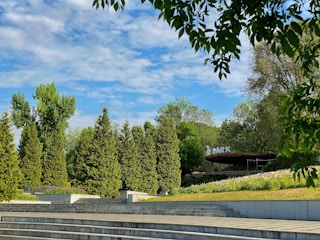 An outdoor scene featuring a landscaped park area with multiple levels of stair-like concrete terraces. Tall, dense evergreen trees and other lush greenery populate the area, with a partly cloudy blue sky above. A section of a sign reading 'PARK' is visible in the background, partially obscured by the foliage.