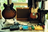 Two electric guitars and an amplifier are situated on a wooden floor in a room filled with natural light. The guitars are positioned to the left and right of the amplifier. Multiple guitar effect pedals are neatly arranged on the floor in front of the guitars, and a bookshelf filled with books is visible on the right.