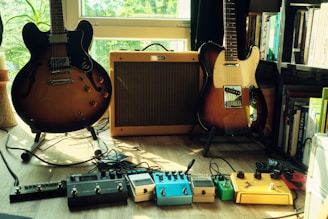 Two electric guitars and an amplifier are situated on a wooden floor in a room filled with natural light. The guitars are positioned to the left and right of the amplifier. Multiple guitar effect pedals are neatly arranged on the floor in front of the guitars, and a bookshelf filled with books is visible on the right.