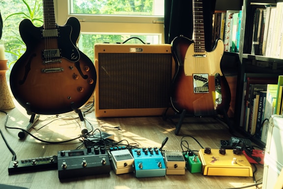 Two electric guitars and an amplifier are situated on a wooden floor in a room filled with natural light. The guitars are positioned to the left and right of the amplifier. Multiple guitar effect pedals are neatly arranged on the floor in front of the guitars, and a bookshelf filled with books is visible on the right.