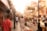 A bustling street scene featuring numerous people walking and shopping. The street is lined with various shops, displaying colorful textiles and items for sale. The architecture showcases traditional designs with ornate details. There is a cart being pushed and a street sign advertising tour packages.